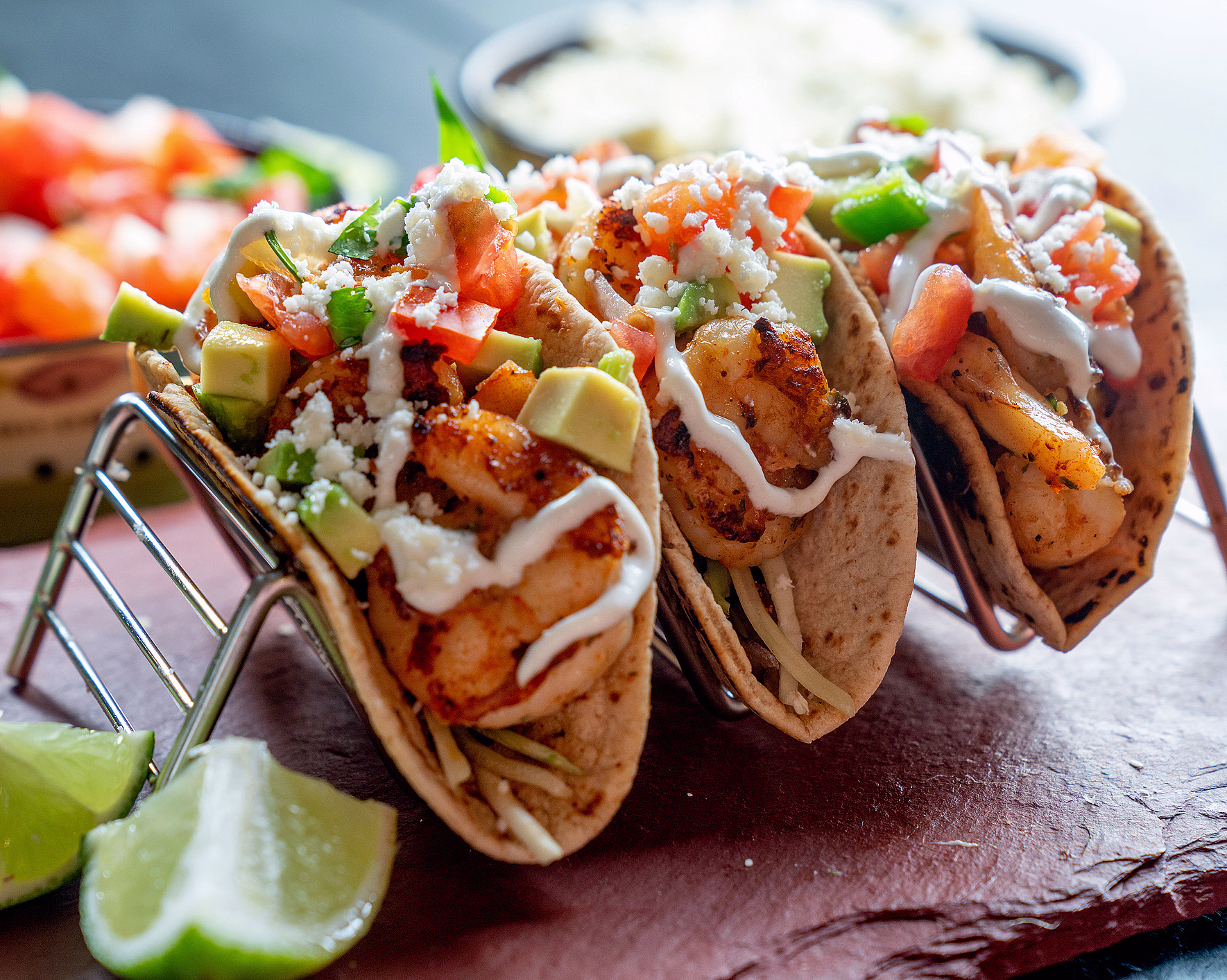 Tre tacoer i metallstativ med grillet fyll, avokado, tomater, hvit saus og limebåter