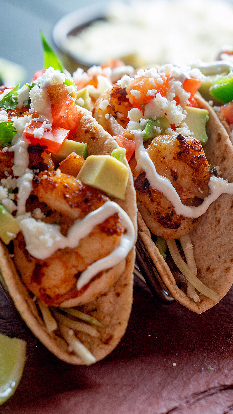 Tre tacoer i metallstativ med grillet fyll, avokado, tomater, hvit saus og limebåter