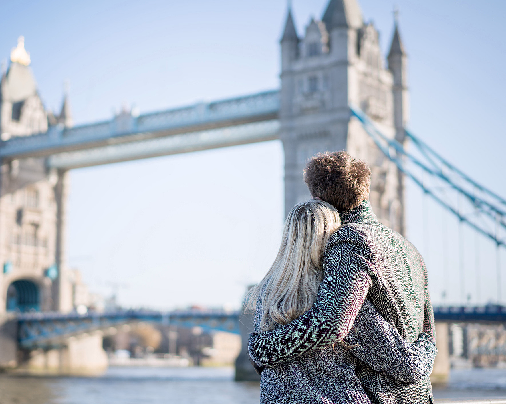 Par med ryggen til ser på Tower Bridge under en klar blå himmel