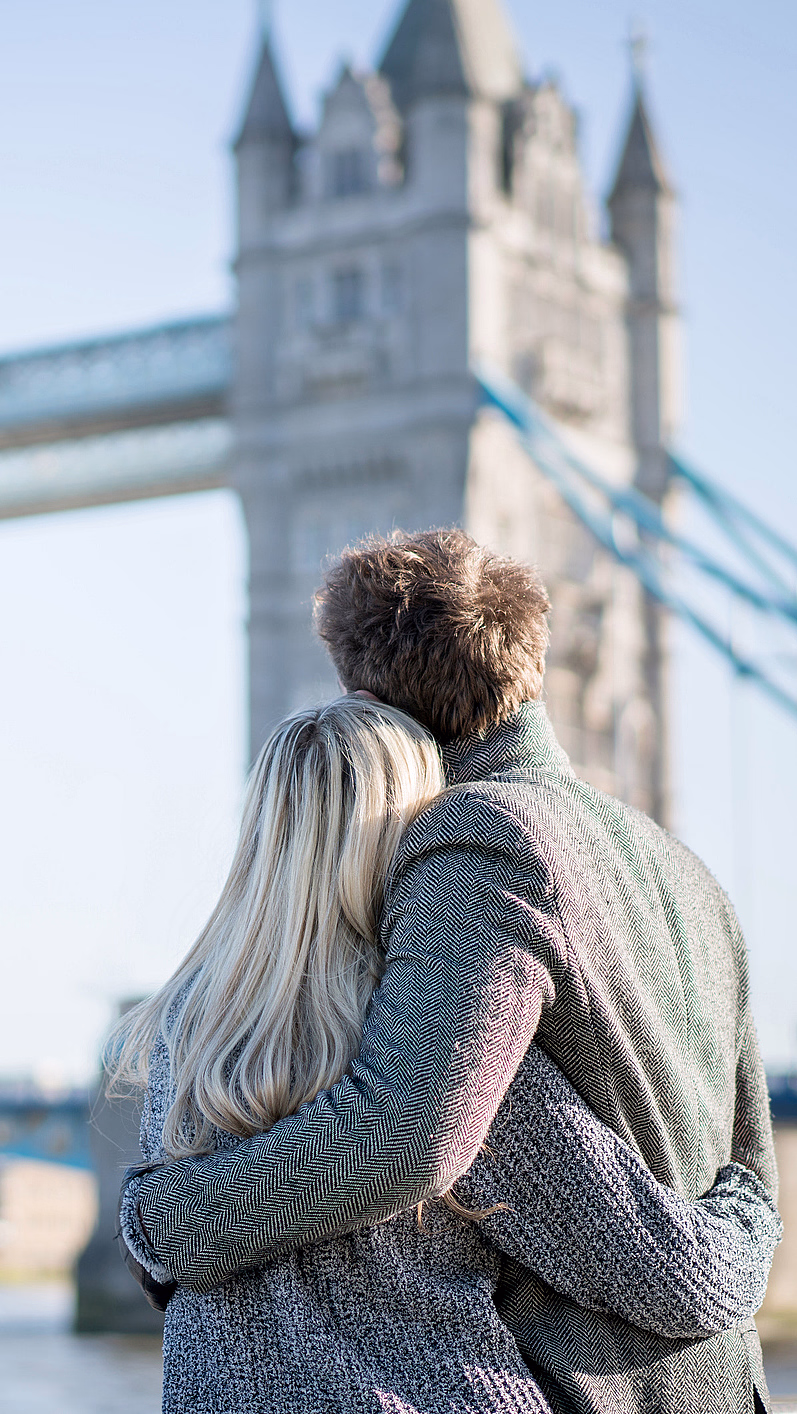 Par med ryggen til ser på Tower Bridge under en klar blå himmel