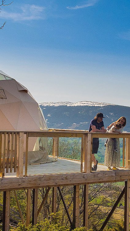 Beige geodesisk kuppel på treterrasse med to personer og utsikt mot åser og snødekte fjell