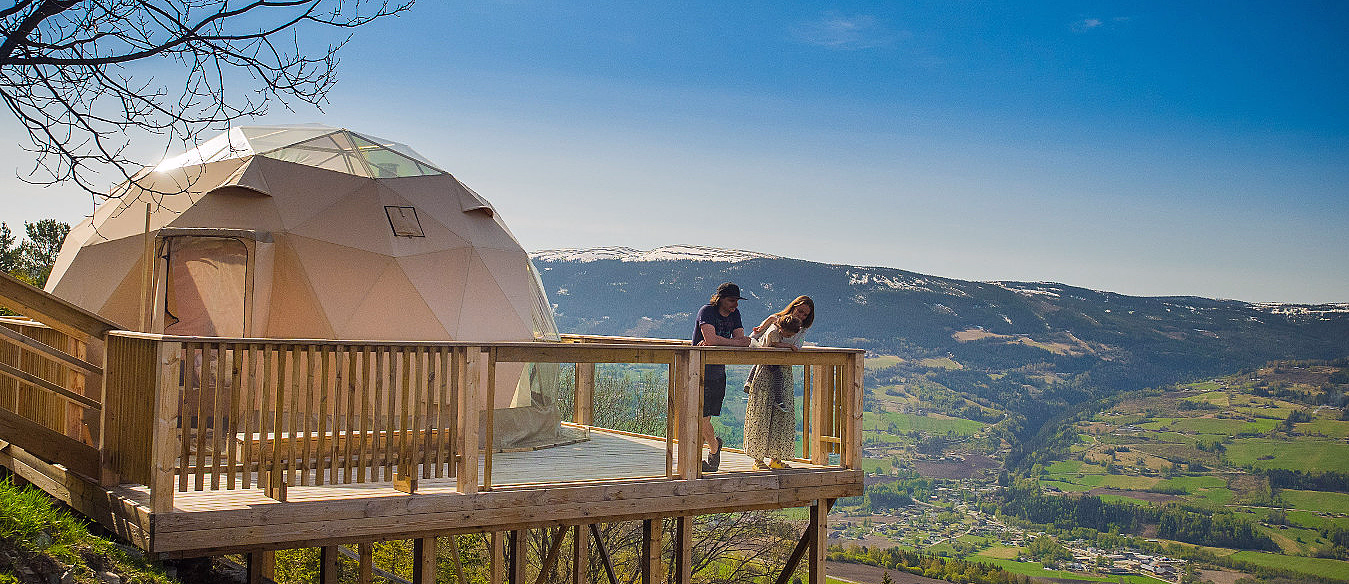 Beige geodesisk kuppel på treterrasse med to personer og utsikt mot åser og snødekte fjell