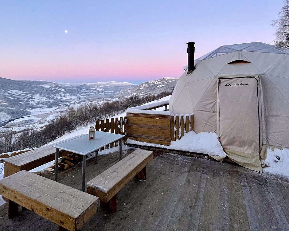 Hvit Arctic Dome på treterrasse i snødekt fjellandskap under rosa og blå kveldshimmel