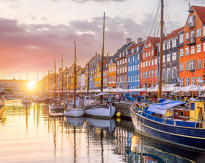 Fargerike bygninger langs en kanal med båter under en rosa og oransje solnedgang