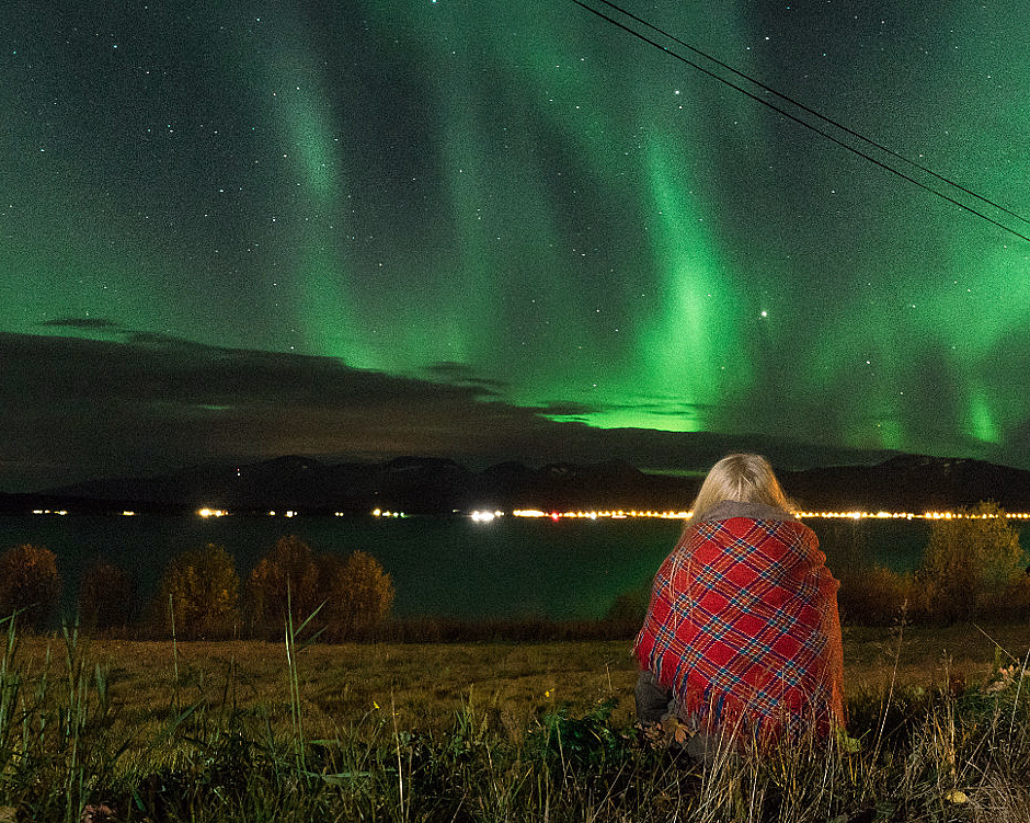 Person i rødrutete pledd sitter på gresset under grønt nordlys over fjell og vann