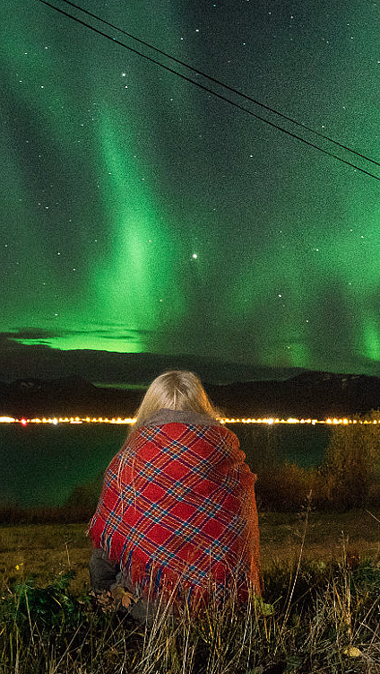 Person i rødrutete pledd sitter på gresset under grønt nordlys over fjell og vann