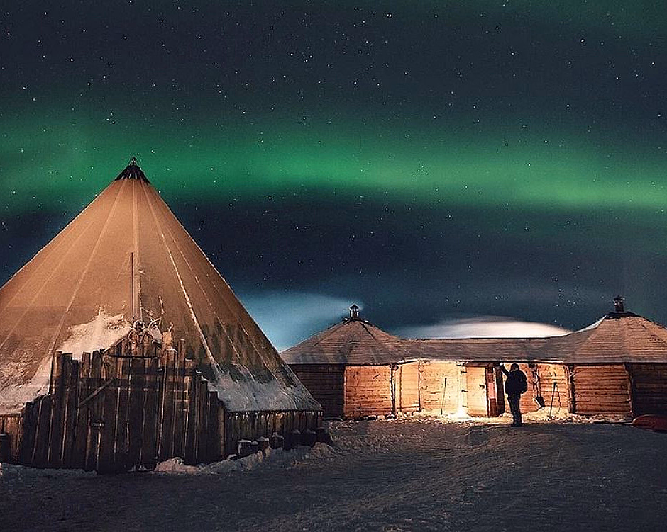 Lavvo og tømmerhytte i snødekt landskap under grønt nordlys og stjerner