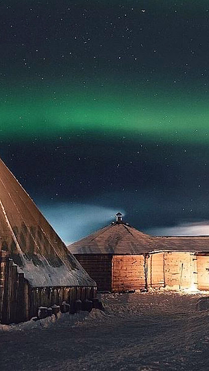 Lavvo og tømmerhytte i snødekt landskap under grønt nordlys og stjerner