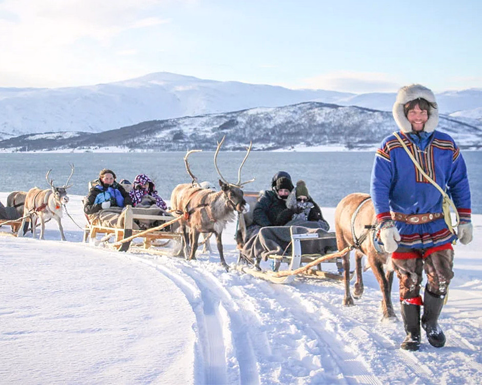 Person som leder rein som trekker sleder med folk i et snødekt fjellandskap