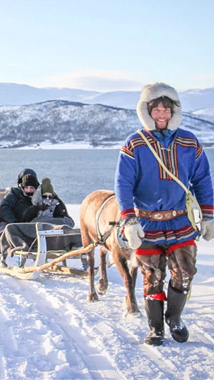 Person som leder rein som trekker sleder med folk i et snødekt fjellandskap