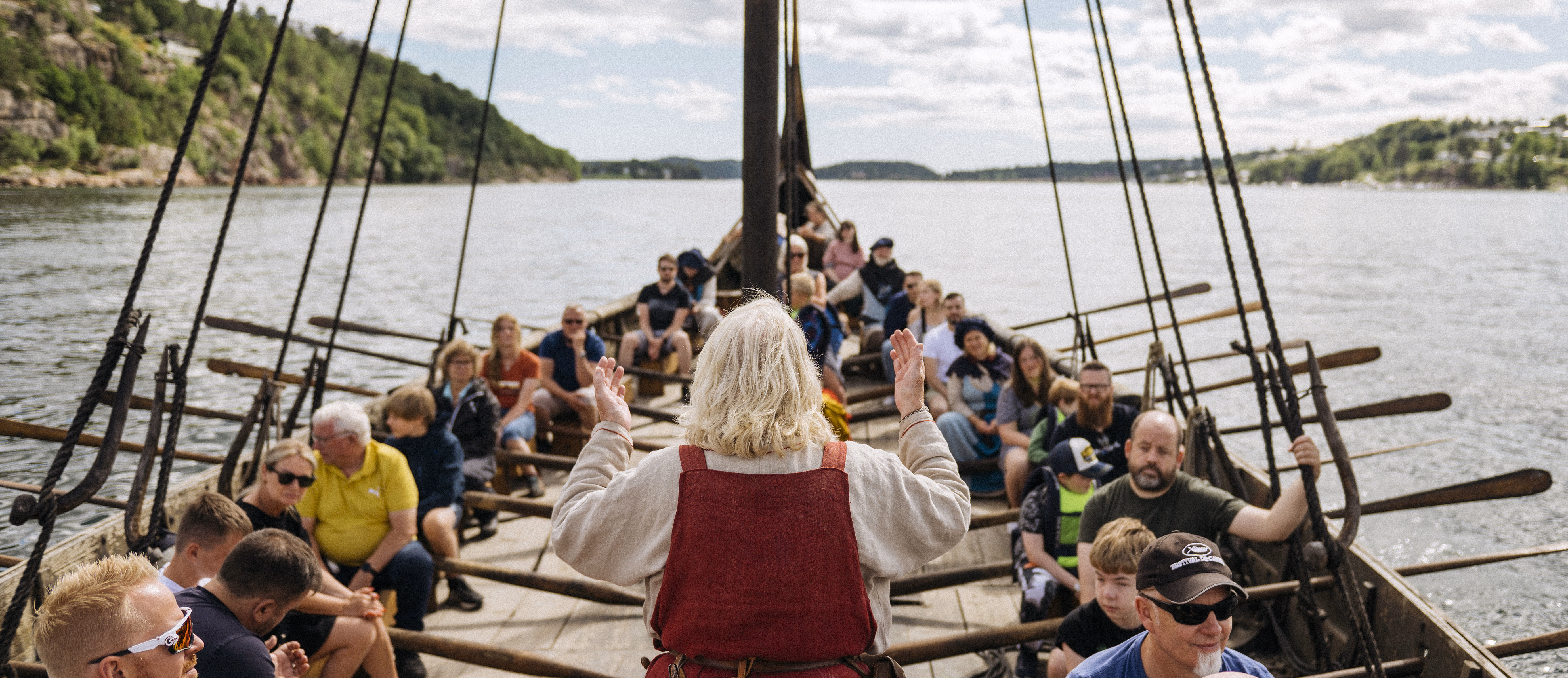 Person med hvitt hår står foran en gruppe i en trebåt med årer på vannet