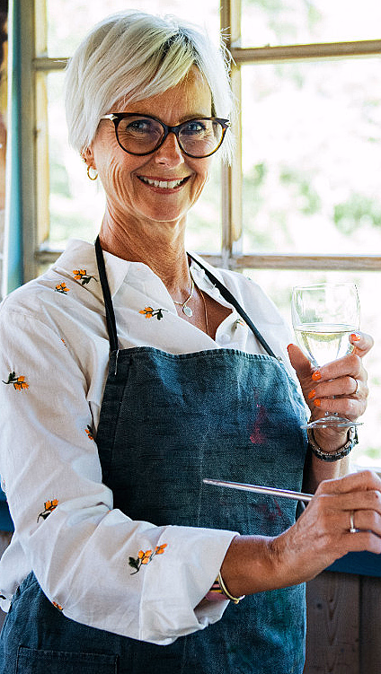 Smilende kvinne med forkle som maler på et staffeli og holder et glass vin