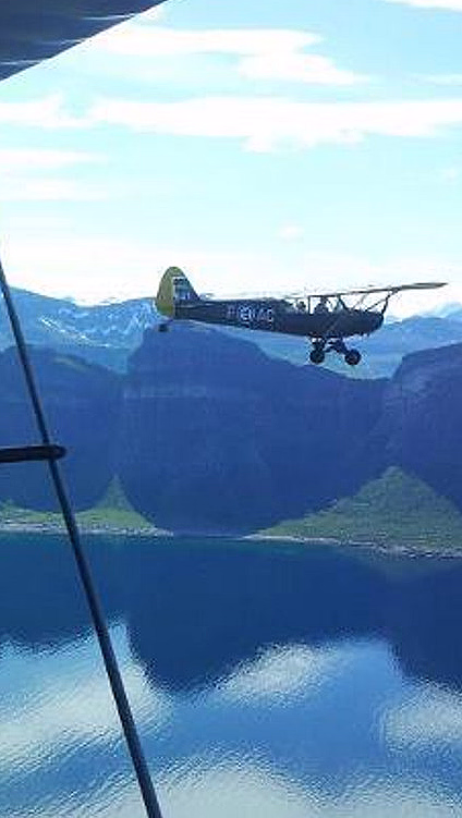 To småfly i luften over en fjord og fjell sett fra vingen av et biplan