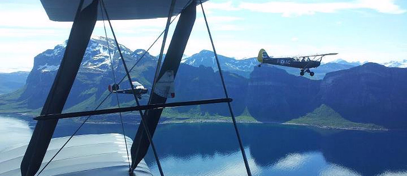 To småfly i luften over en fjord og fjell sett fra vingen av et biplan