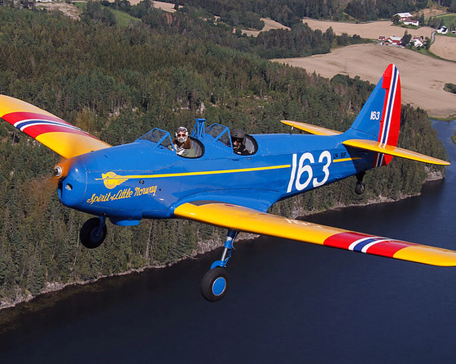 Blått veteranfly med gule vinger og tallet 163 flyr over skog og elv