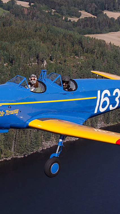 Blått veteranfly med gule vinger og tallet 163 flyr over skog og elv