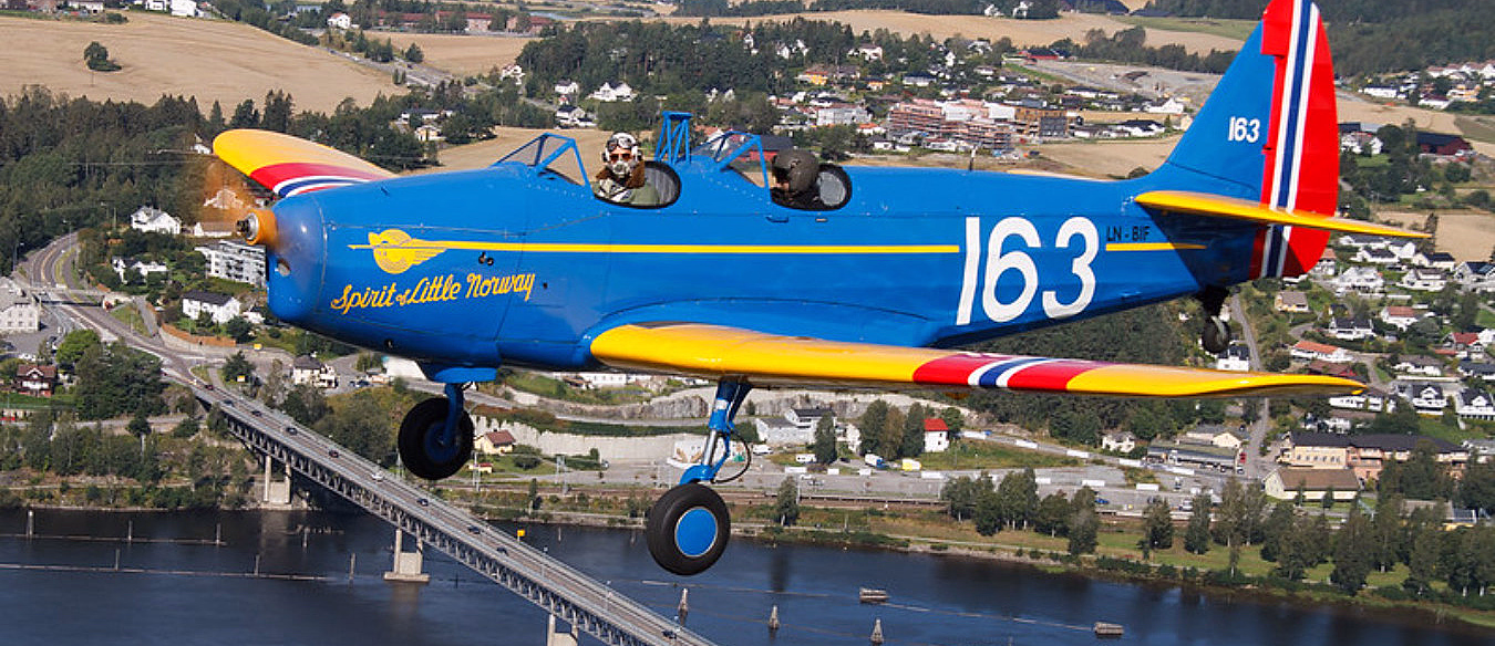 Blått veteranfly merket Spirit of Little Norway 163 flyr over en elv og en bro