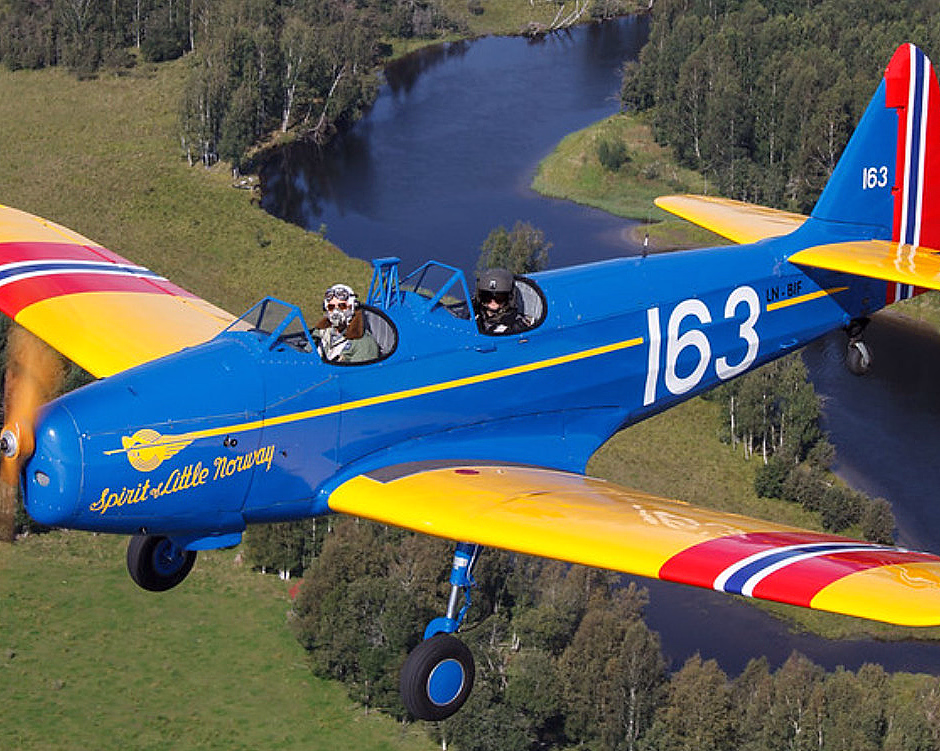 Blått fly med gule vinger og teksten Spirit of Little Norway 163 flyr over skog og elv