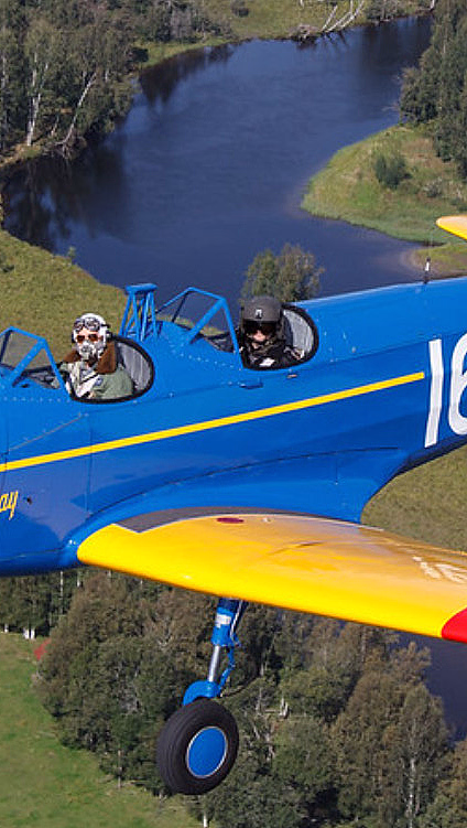 Blått fly med gule vinger og teksten Spirit of Little Norway 163 flyr over skog og elv
