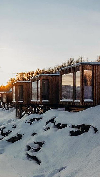 Moderne trehytter på stylter ved en snødekt fjord i solnedgang
