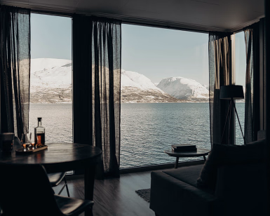 Utsikt fra en moderne hytte mot en fjord og snødekte fjell gjennom store vinduer