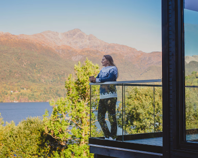 Kvinne i strikkegenser står på en balkong med utsikt over fjell og fjord