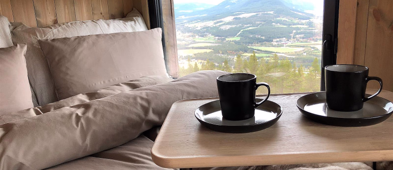 Seng med kaffekopper på et brett foran et vindu med utsikt over fjell og skog
