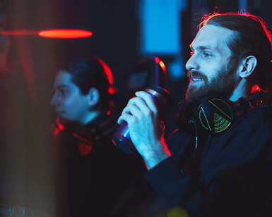 Mann med skjegg og hodetelefoner sitter i en gamingstol og holder en kopp i et rom med neonlys