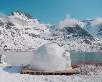 Hvit glassiglo på en treplattform ved vannet med snødekte fjell i bakgrunnen