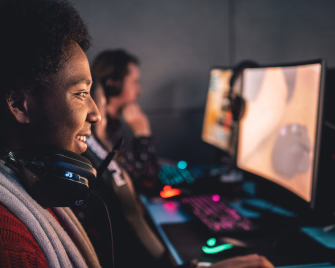 Smilende person med headset foran en dataskjerm i et mørkt rom med gamingutstyr