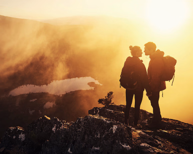 To personer med ryggsekker står på en fjelltopp i solnedgang og ser på hverandre