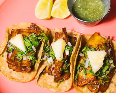 Tre tacos med ananas, løk og koriander servert med lime og grønn saus på en rosa tallerken