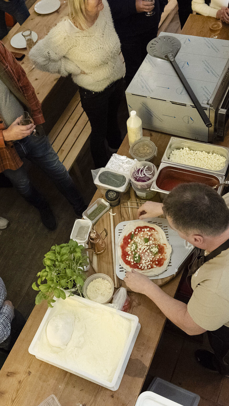 En gruppe mennesker ser på en mann som tilbereder en pizza på et kjøkkenbord