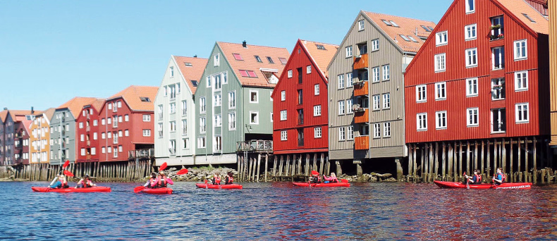 Folk i røde kajakker på vannet foran fargerike bryggehus i tre