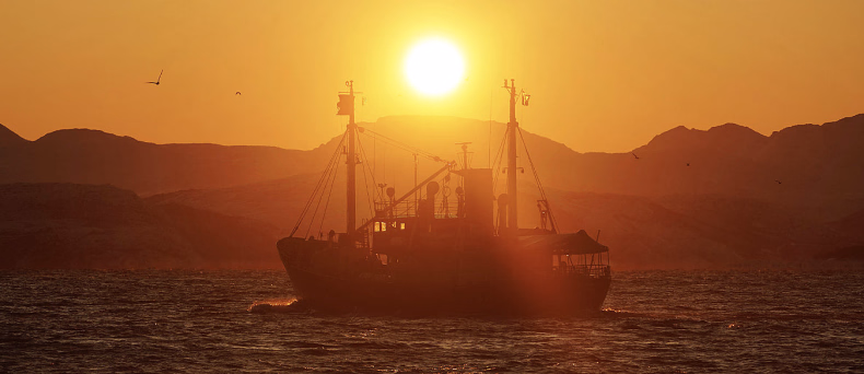 Silhuett av en båt på havet i solnedgang med fjell i bakgrunnen