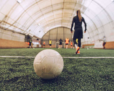 Nærbilde av en fotball på en kunstgressbane i en innendørshall med spillere i bakgrunnen