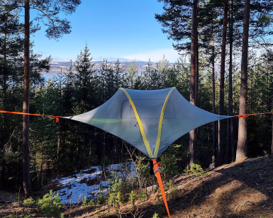 Grått svevetelt spent opp mellom trær i en skog med utsikt over vannet
