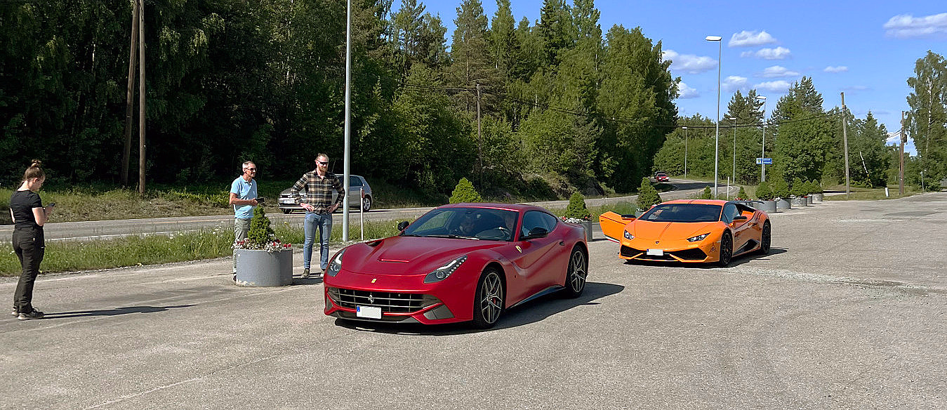 Rød og oransje sportsbil parkert på en asfaltert plass med skog i bakgrunnen