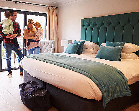 En familie på fire står i et hotellrom med en stor seng og bagasje på gulvet