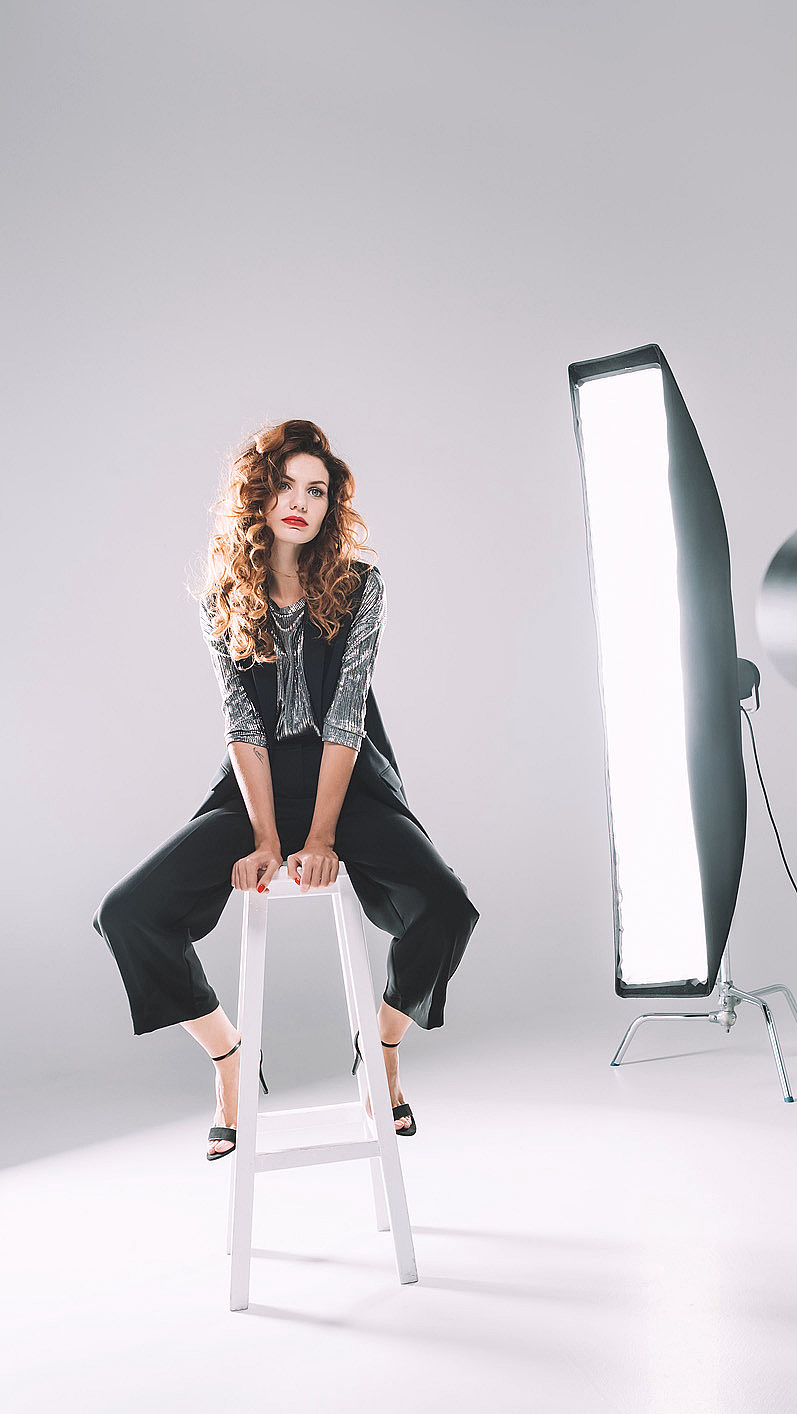 Kvinne med krøllete hår på hvit krakk i et fotostudio med lamper og kamera