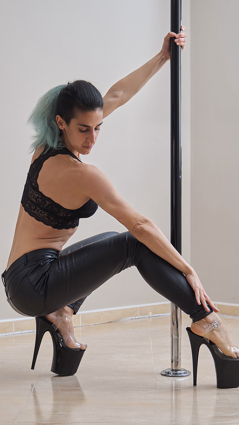 Person med lyseblått hår poserer ved en dansepole i sort blondetopp og platåsko