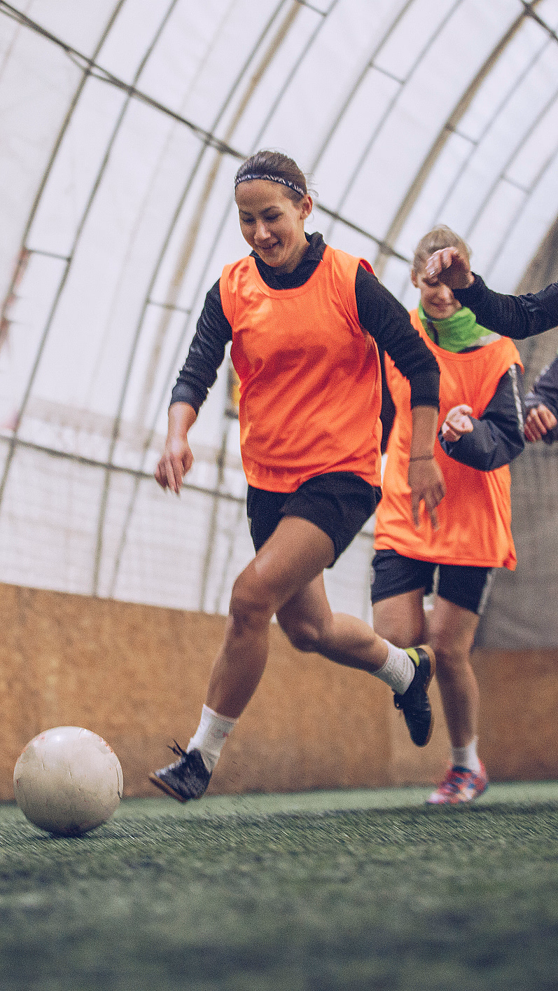 Fire personer i gule og oransje vester spiller fotball på grønt kunstgress innendørs