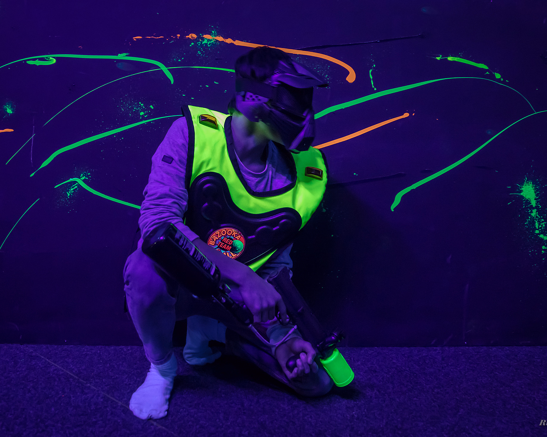 Person i neongrønn vest og maske med paintballmarkør i et mørkt rom med neonflekker