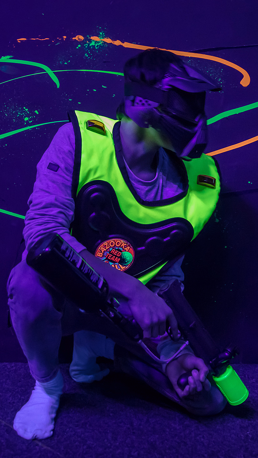 Person i neongrønn vest og maske med paintballmarkør i et mørkt rom med neonflekker