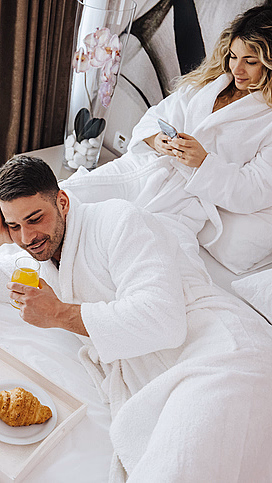 Mann og kvinne i hvite badekåper på en seng med juice, en croissant og en mobil