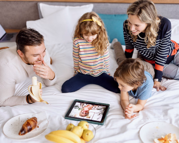 Familie på fire i en seng ser på et nettbrett med frukt og bakverk