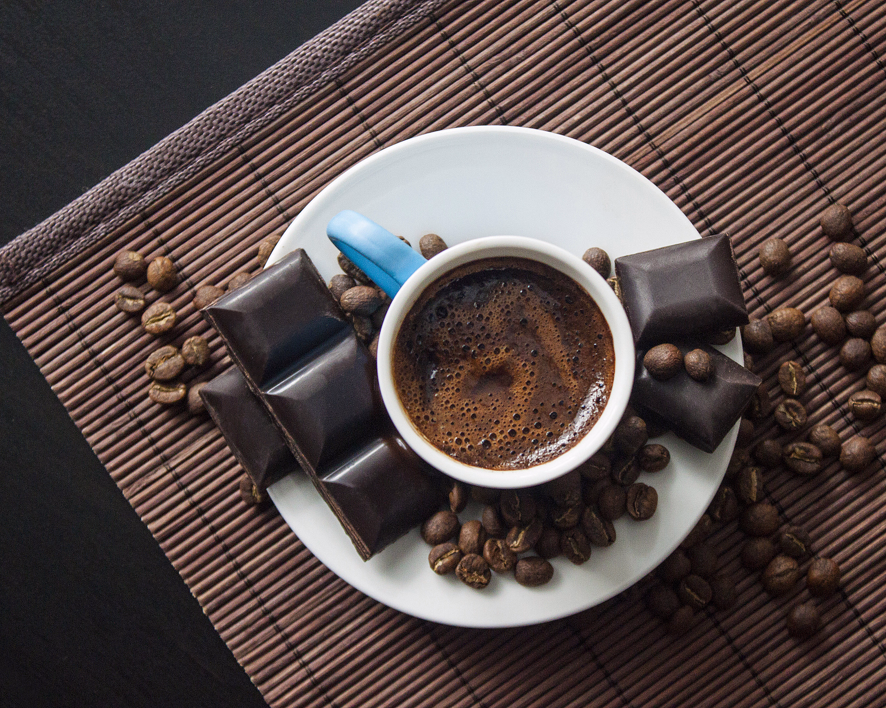 Hvit kaffekopp med skum, kaffebønner og mørk sjokolade på en bambusmatte