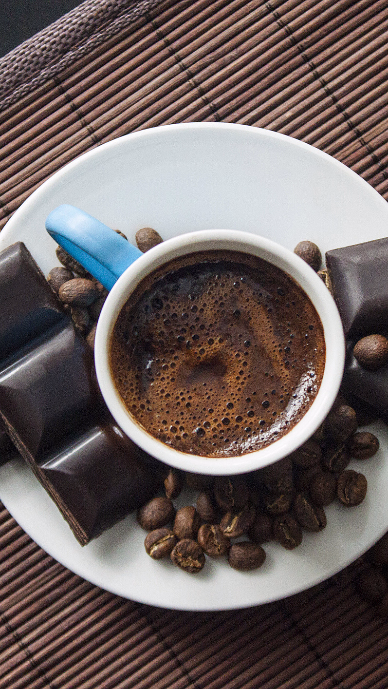 Hvit kaffekopp med skum, kaffebønner og mørk sjokolade på en bambusmatte