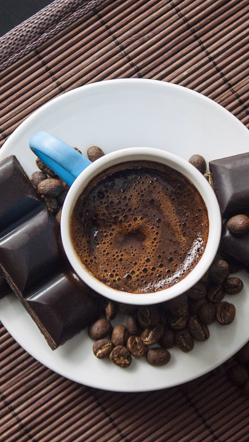 Hvit kaffekopp med skum, kaffebønner og mørk sjokolade på en bambusmatte