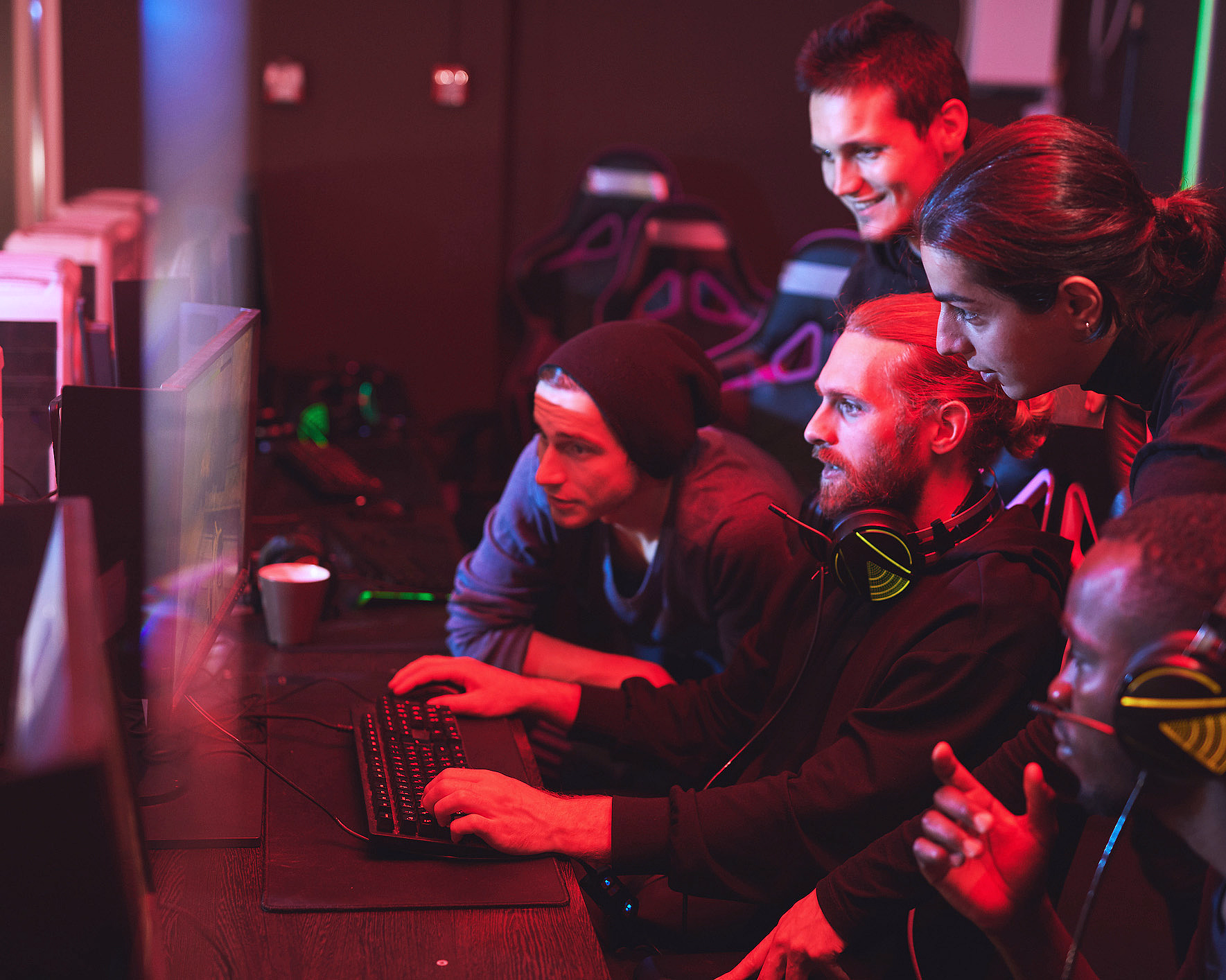 Fire personer med headset ser på en dataskjerm i et rom med rødt lys og gamingstoler